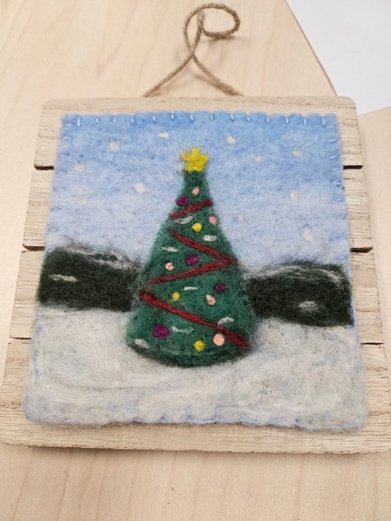 A needle felted Christmas tree decorated with small coloured baubles and a yellow star, standing in snow under a pale blue sky.