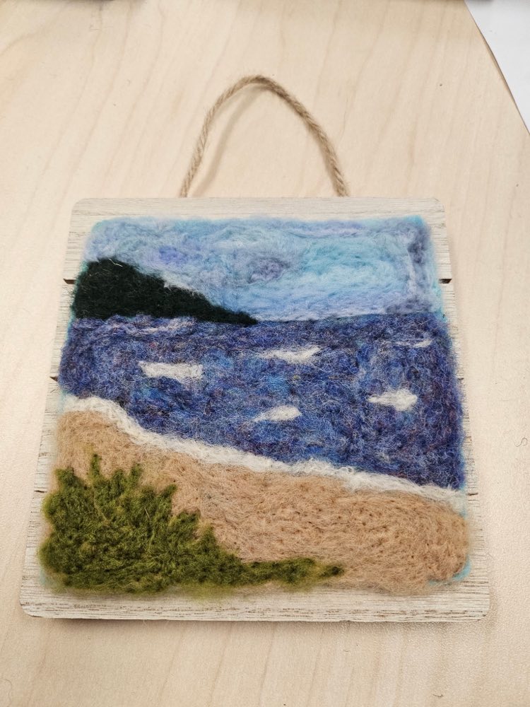 A needle felted coastal scene mounted on pale wood: a dark green headland above deep blue water flecked with white, a pale sand beach, and a mossy green shrub in the foreground.