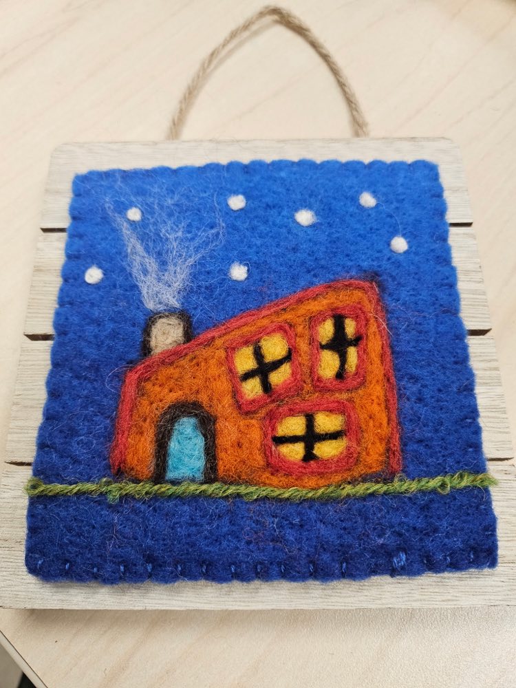 A needle felted orange house at night with lit yellow windows and pale smoke curling from the chimney, against a deep blue snowy sky.