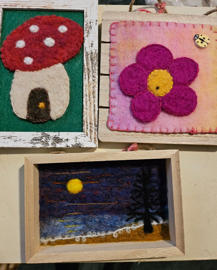 Three of Liz's framed needle felted pieces: a red-capped mushroom house with a small door, a magenta flower blanket stitched onto pink felt with a ladybug button beside it, and a moonlit scene with a black tree above gold sand.