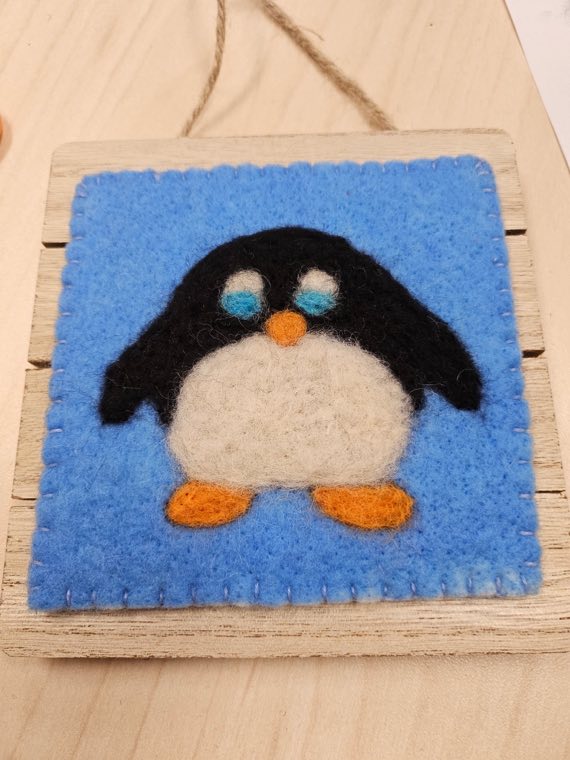 A needle felted penguin with an orange beak and orange feet on a bright blue background, blanket stitched around the edge and mounted on a wooden board.