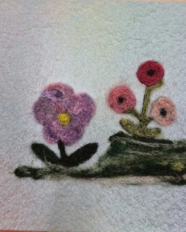 Needle felted purple, red and pink flowers growing from a dark green mound on pale felt.