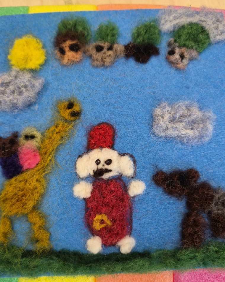 A busy needle felted scene on bright blue: a white dog in a red outfit, a tall yellow giraffe carrying a little bear, a brown dog, clouds, a yellow sun and a row of small faces across the top.