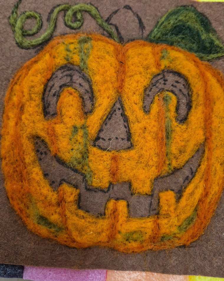 A needle felted jack-o'-lantern face in bright orange with a curling green vine and leaf, on brown felt.