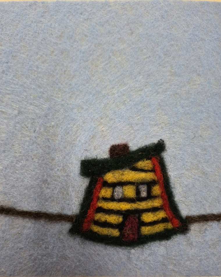 A needle felted log cabin with yellow and red stripes and a green roof, sitting on a thin brown line on pale grey felt.