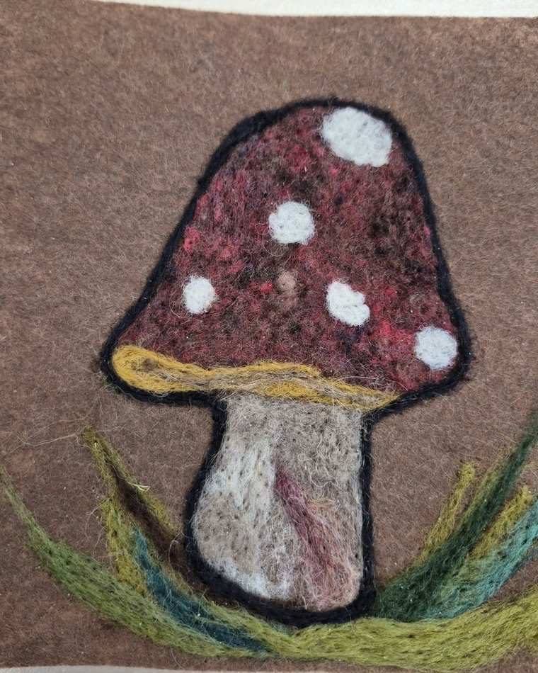 A needle felted red toadstool with white spots and a pale stem, standing in tufts of green and teal grass on brown felt.