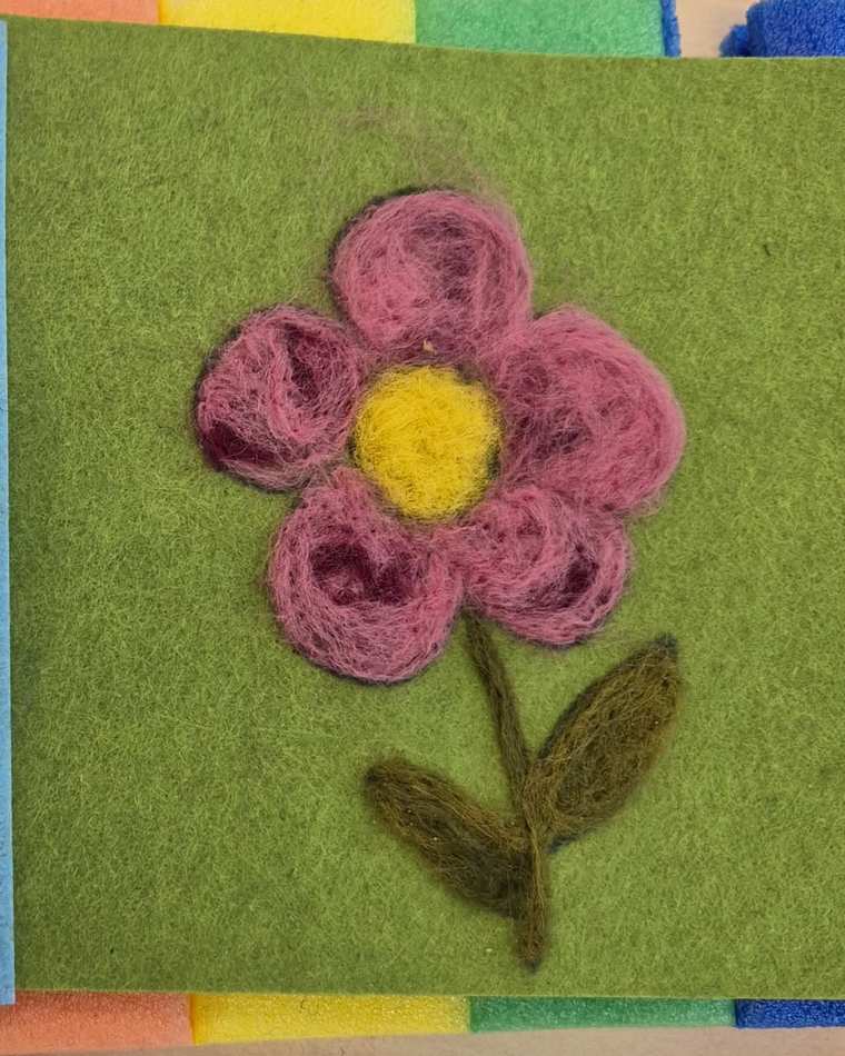 A needle felted pink flower with a yellow centre and two olive leaves, on green felt.