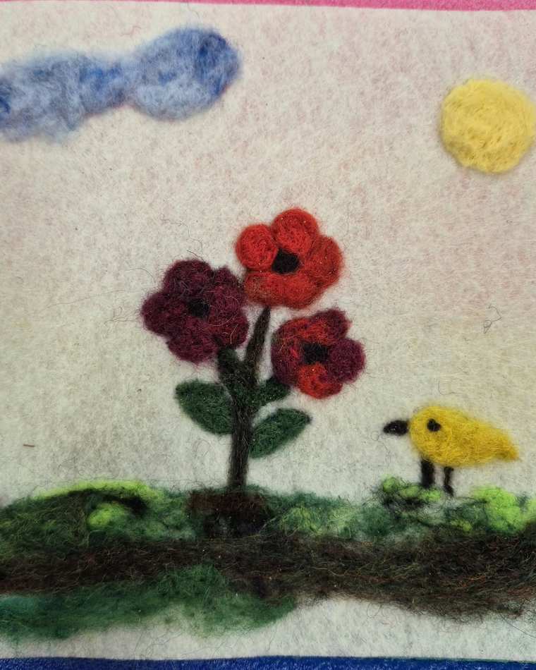 A needle felted scene of red and deep red poppies on a leafy stem beside a small yellow bird on green grass, under a blue cloud and a yellow sun.