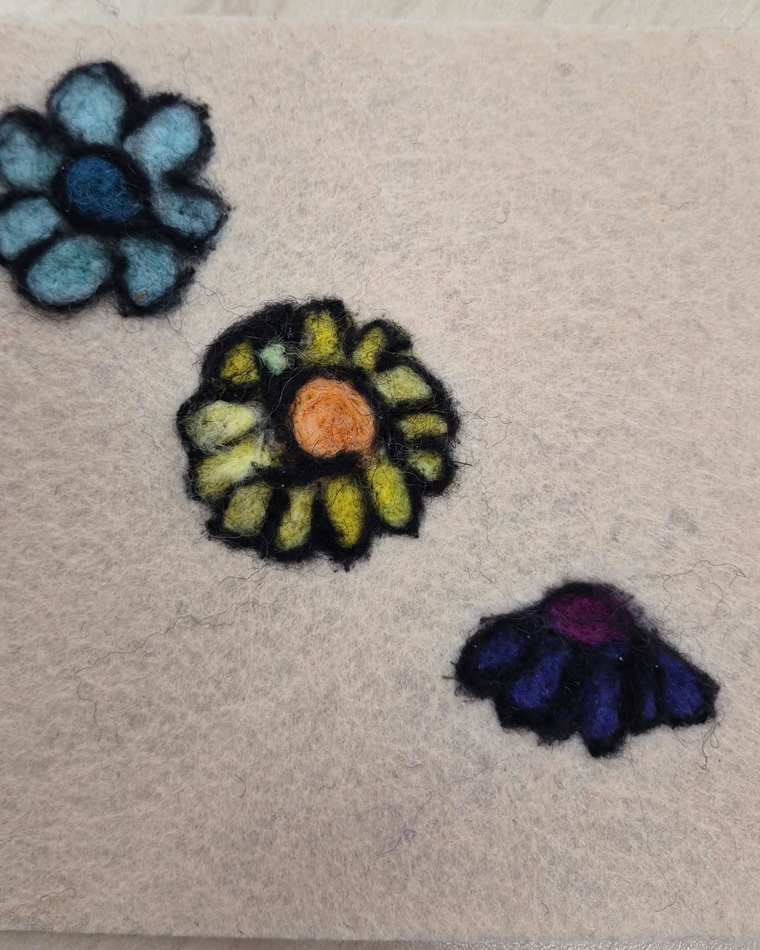 Three needle felted flowers outlined in black, blue, yellow and purple, scattered across pale felt.