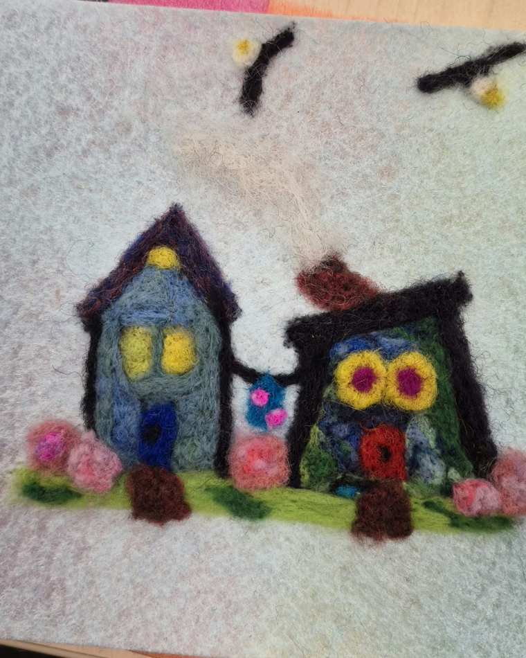 Two needle felted houses side by side, one blue and one green with round yellow windows, with pink flowers along the grass and two birds overhead.