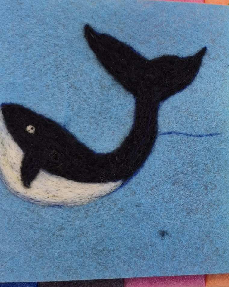 A needle felted black and white whale with its tail raised, on pale blue felt.