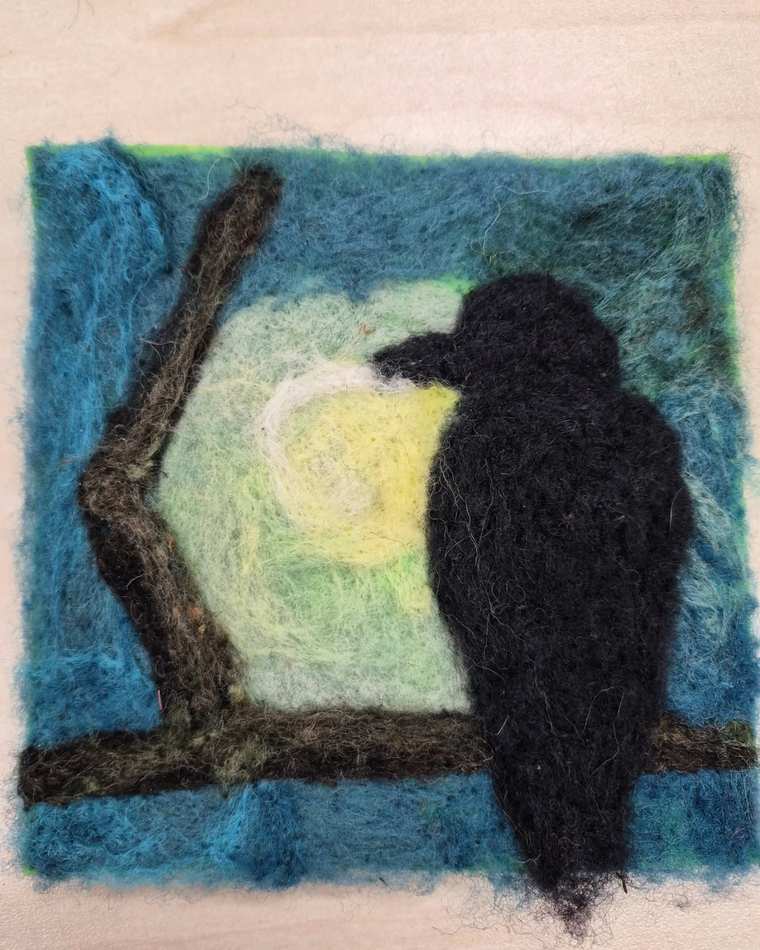 A needle felted black crow perched on a bare brown branch in front of a glowing pale yellow moon, against a deep teal sky.