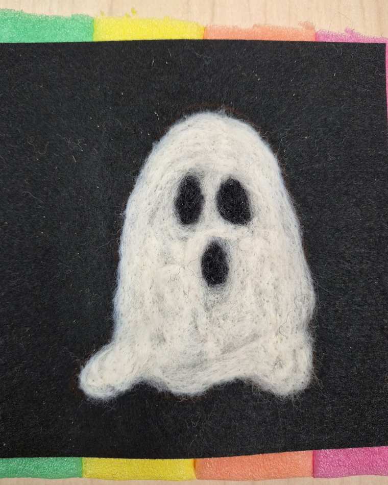 A needle felted white ghost with dark oval eyes and an open mouth on black felt.