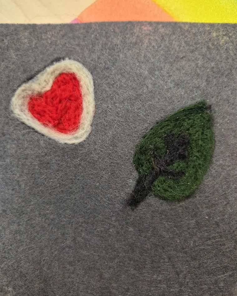 A needle felted red heart outlined in cream and a green leaf with a dark stem, side by side on grey felt.