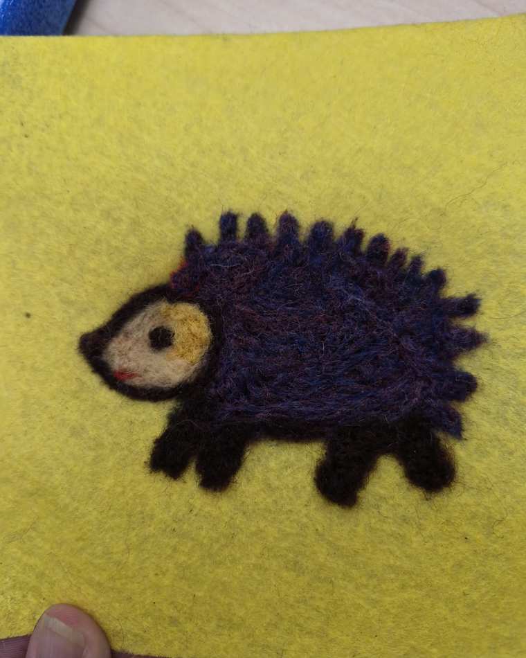 A needle felted hedgehog with dark purple spines, a tan face and a small pink nose, walking across bright yellow felt.