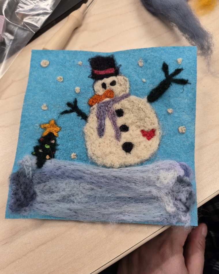 A needle felted snowman in a black top hat, orange bow tie and purple scarf, with stick arms and a small red heart, beside a little star-topped tree on a snowbank against a light blue sky dotted with snow.