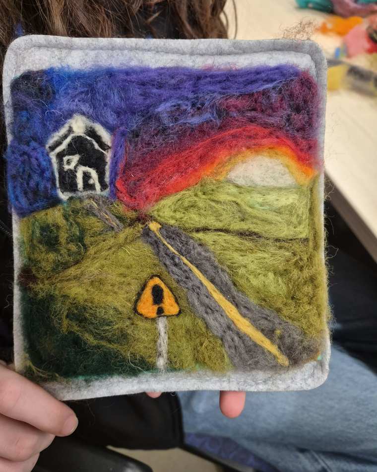 A needle felted landscape: a winding grey road with a yellow centre line runs through green fields toward a small dark house on a hill, under a sunset sky of purple, red and orange, with a yellow warning sign by the roadside. Held up in two hands.