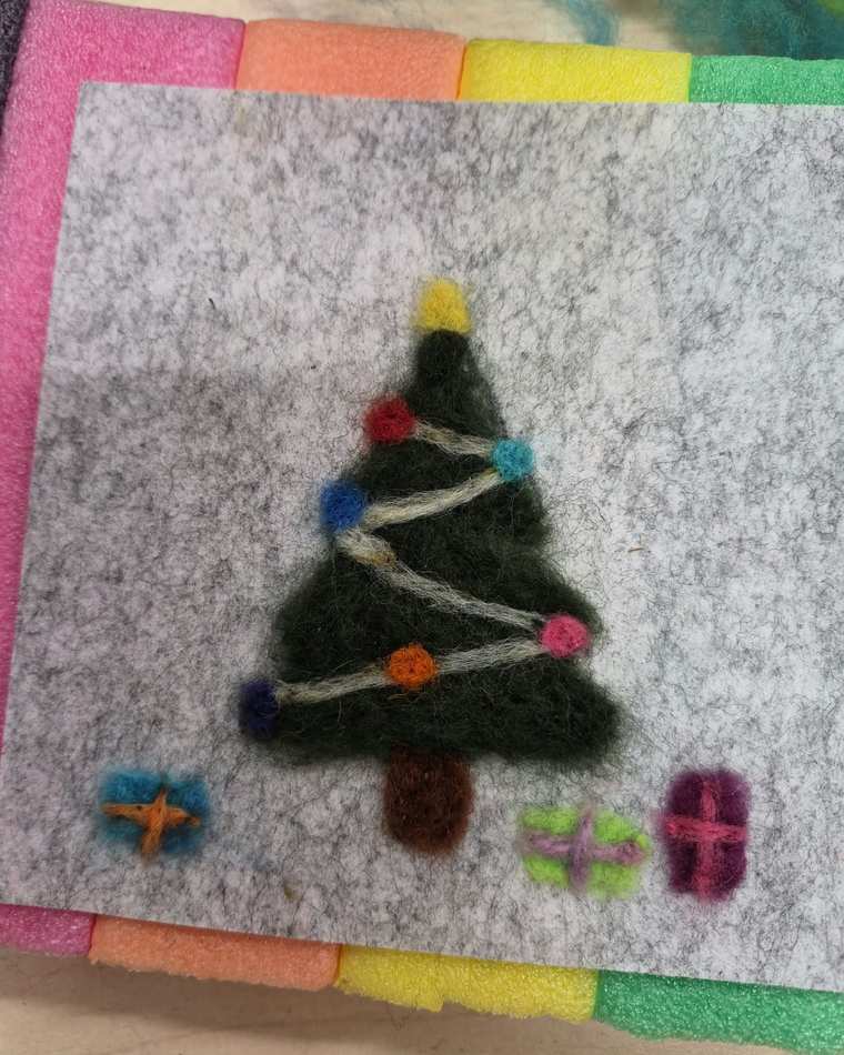 A needle felted Christmas tree strung with coloured baubles and a yellow star on its tip, with three small wrapped presents beneath it, on pale grey felt.