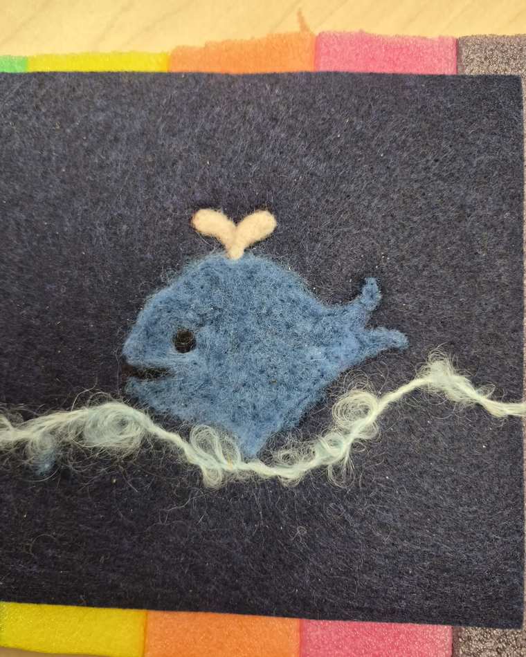 A needle felted blue whale with a cream spout, swimming above a curly wave of pale green wool on dark charcoal felt.
