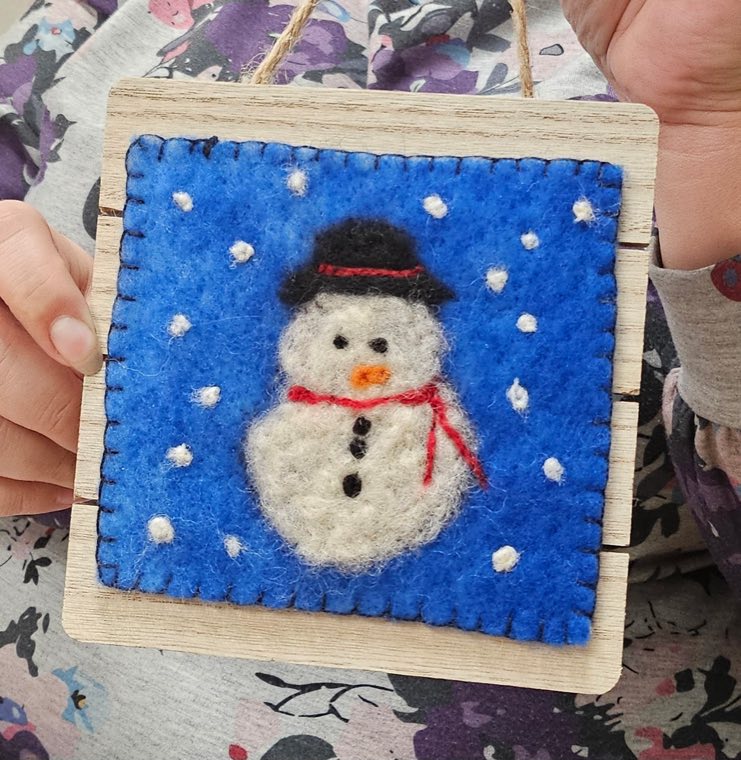 A needle felted snowman in a black top hat and red scarf on a bright blue background dotted with white snow, held up in two hands.