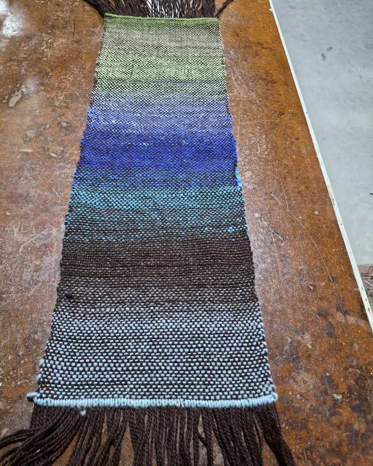 A handwoven sample shading from pale green through deep blue and teal into dark brown and a grey-blue band, with a long brown fringe.