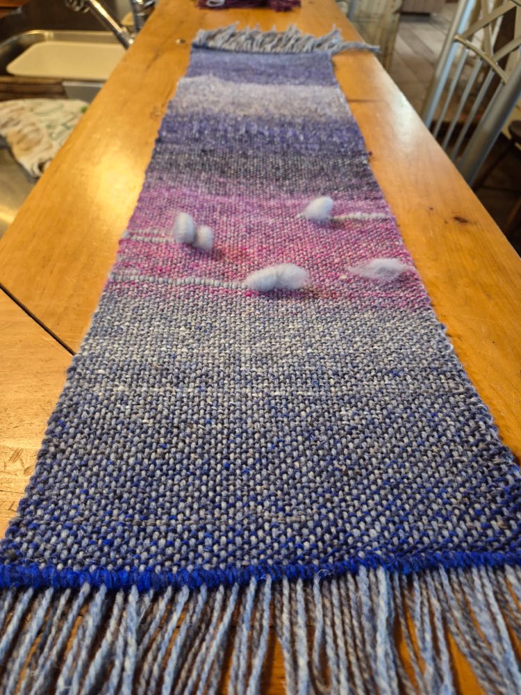 A handwoven sample in blues and violets laid out along a wooden table, with a band of soft white tufts worked into the middle and a long fringe at the near end.