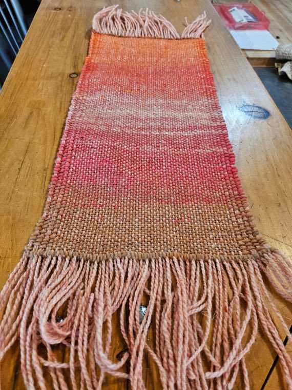 A handwoven sample shading from orange through coral and pink into sandy brown, with a pale pink fringe.