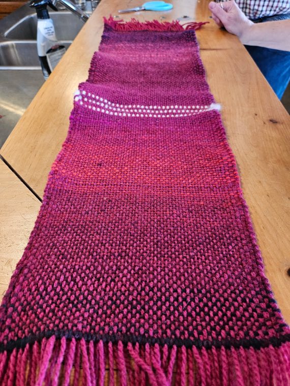 A handwoven sample in magenta and crimson with a row of small white dots woven in a line across it.