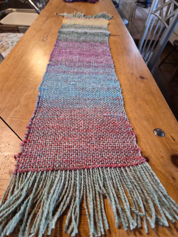 A handwoven sample shading from cream through teal into dusty pink, finished with a long grey-green fringe.