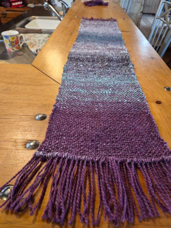 A handwoven sample in deep plum with a band of soft teal and grey through the centre, finished with a thick purple fringe.